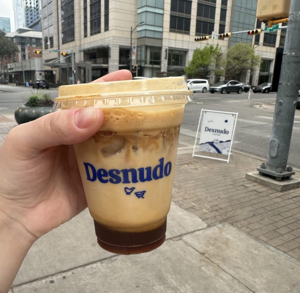 Person holding up iced coffee from Desnudo coffee in front of city street